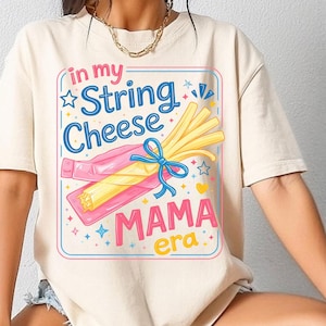 Könnte beinhalten: Cremefarbenes T-Shirt mit farbenfroher Grafik. Die Grafik zeigt den Text "in my String Cheese MAMA era" mit Illustrationen von Käsefäden und einer Schleife. Das Design ist von einem hellblauen und rosa Rahmen umgeben. Ein lässiges Kleidungsstück.