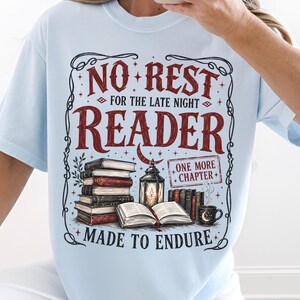 以下が含まれることがあります： NO REST FOR THE LATE NIGHT READERおよびMADE TO ENDUREのテキストがプリントされたライトブルーのTシャツ。デザインには、本の山、開いた本、火のついたランタン、マグカップ、ONE MORE CHAPTERのテキストが含まれています。