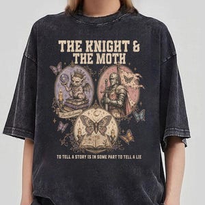 Könnte beinhalten: Ein anthrazitfarbenes T-Shirt in Acid-Wash-Optik mit einem Grafikdruck, der den Text THE KNIGHT & THE MOTH und TO TELL A STORY IS IN SOME PART TO TELL A LIE zeigt. Das Design enthält Illustrationen eines Wasserspeiers, eines Ritters und eines Falters.