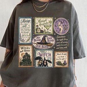 Op de afbeelding: Een antracietgrijs T-shirt met korte mouwen en een collage van rechthoekige en ronde afbeeldingen in vintage-stijl. De prints bevatten teksten zoals Always salt, Midnight Margaritas, My Blood Our Blood, Witch's House en There's A Little Witch In All Of Us.