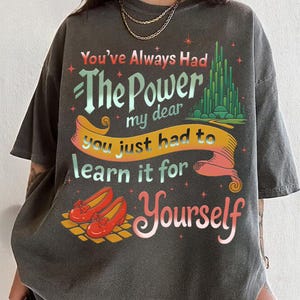 May include: Dark gray t-shirt with the text "You've Always Had The Power my dear you just had to learn it for Yourself." The design includes red shoes and a green city illustration.