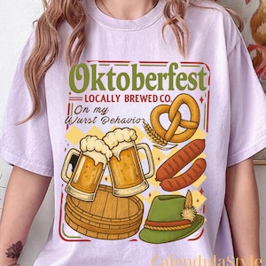 May include: A lilac t-shirt featuring an Oktoberfest design. The design includes the word "Oktoberfest", beer mugs, a pretzel, sausages, and a green hat. Text reads "Locally Brewed Co." and "On my Wurst Behavior."