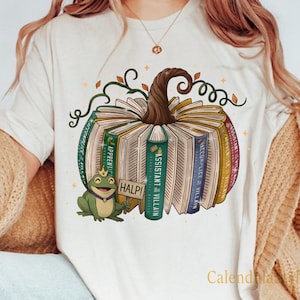 Puede incluir: Camiseta color crema con un diseño de calabaza hecha de libros, un tallo marrón y enredaderas verdes. Una rana verde con una corona y la palabra "HALP!" está a la izquierda. La camiseta tiene el texto "Calendula Life" en la parte inferior derecha.
