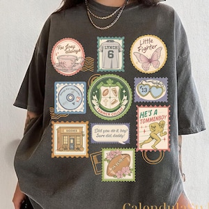 Puede incluir: Camiseta gris oscuro con varios diseños de sellos postales. Los sellos incluyen una taza de té, una camiseta con el número 6, una mariposa, un CD, un escudo escolar, un corazón, una tienda, un balón de fútbol y una figura corriendo. Textos: "For Joey always", "Little Fighter" y "He's a Tommenboy".