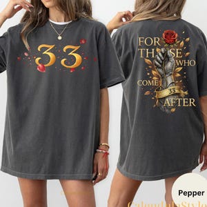 Puede incluir: Camiseta gris oscuro con el número "33" en dorado en la parte delantera. La parte trasera de la camiseta presenta una mano sosteniendo una rosa, con el texto "FOR THOSE WHO COME 33 AFTER" en dorado.