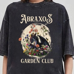 以下が含まれることがあります： 黒いTシャツに、ヴィンテージスタイルの黒いドラゴンが花のある庭にいるグラフィックと「Abraxo's Garden Club」の文字がプリントされています。