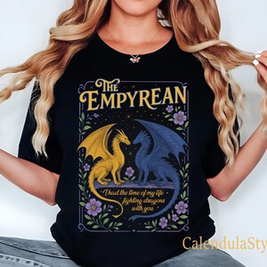 Może przedstawiać: Czarna koszulka z nadrukiem z napisem "The Empyrean" i dwoma smokami, jednym złotym i jednym niebieskim. Projekt zawiera fioletowe kwiaty i cytat "I had the time of my life fighting dragons with you."