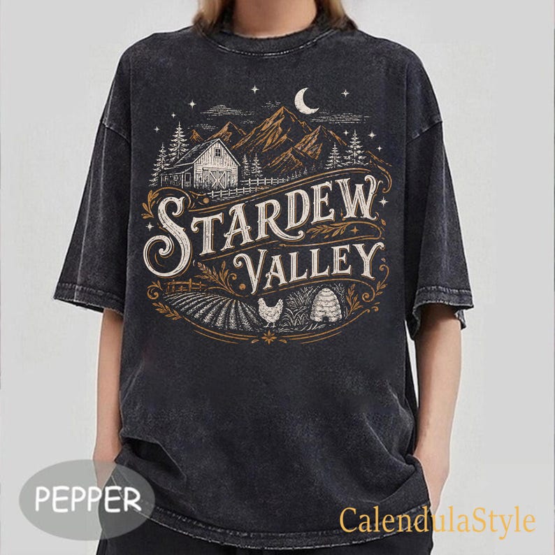 Puede incluir: Camiseta gris oscuro con un dise&ntilde;o vintage que dice "Stardew Valley". El dise&ntilde;o incluye un granero, monta&ntilde;as, &aacute;rboles, una luna creciente y un campo con una gallina y una colmena. La camiseta tiene un aspecto ligeramente descolorido.