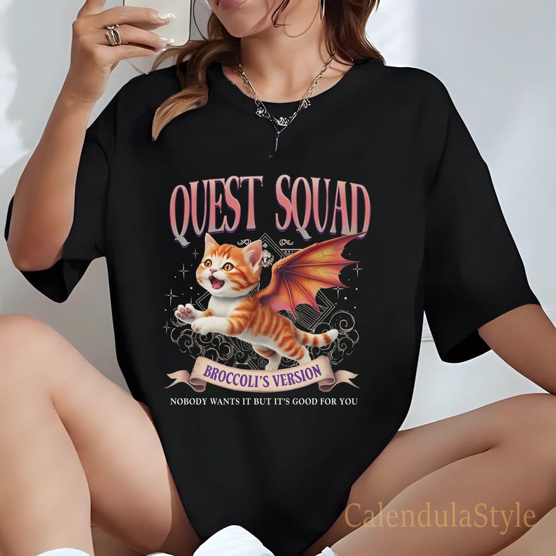 Może przedstawiać: Czarny T-shirt z grafiką pomarańczowego kreskowego kota z skrzydłami. Tekst "Quest Squad" znajduje się nad kotem, a "Broccoli's Version" pod kotem. Tekst "Nobody wants it but it's good for you" znajduje się pod banerem.