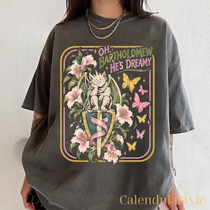May include: A charcoal grey short-sleeve t-shirt featuring a graphic print of a gargoyle perched on a sword, surrounded by pink lilies and yellow and pink butterflies. The text reads, "OH, BARTHOLOMEW HE'S DREAMY" in a stylised, decorative font.