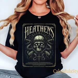 May include: Black t-shirt featuring a detailed graphic with the word "Heathens" above a skull encircled by snakes and a crown of thorns. The design includes text at the bottom and is framed in a decorative border. The shirt has a crew neck.