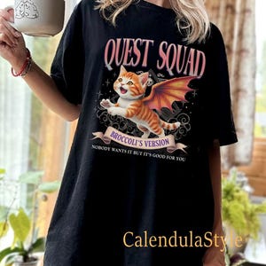 Może przedstawiać: Czarny T-shirt z grafiką przedstawiającą pomarańczowego kota z kresk&oacute;wek z r&oacute;żowymi skrzydłami. Tekst "QUEST SQUAD" znajduje się nad kotem, a "BROCCOLI'S VERSION" pod kotem. Tekst "NOBODY WANTS IT BUT IT'S GOOD FOR YOU" znajduje się pod grafiką.