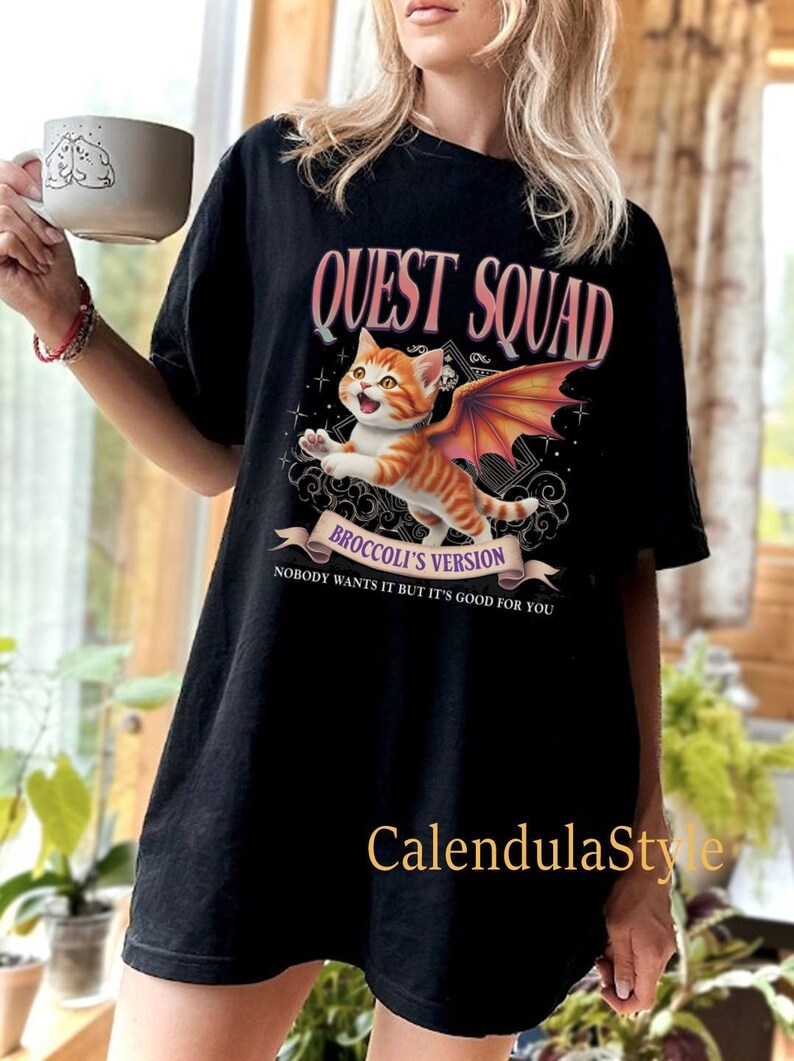 Może przedstawiać: Czarny T-shirt z grafiką przedstawiającą pomarańczowego kota z kresk&oacute;wek z r&oacute;żowymi skrzydłami. Tekst "QUEST SQUAD" znajduje się nad kotem, a "BROCCOLI'S VERSION" pod kotem. Tekst "NOBODY WANTS IT BUT IT'S GOOD FOR YOU" znajduje się pod grafiką.