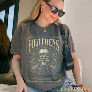 May include: Dark grey t-shirt featuring a skull design with snakes, a cross, and the word "HEATENS" in a vintage style. The design is in shades of green and beige. The shirt is worn by a person wearing sunglasses.