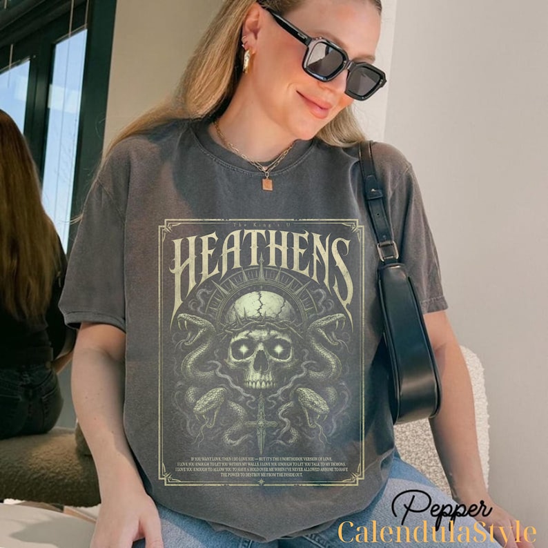 May include: Dark grey t-shirt featuring a skull design with snakes, a cross, and the word "HEATENS" in a vintage style. The design is in shades of green and beige. The shirt is worn by a person wearing sunglasses.