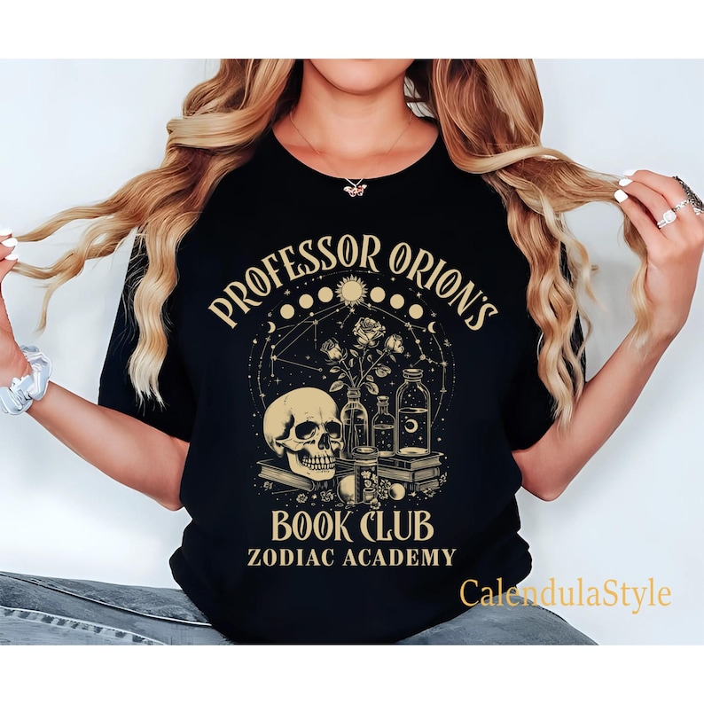 Puede incluir: Camiseta negra con un dise&ntilde;o gr&aacute;fico dorado que presenta una calavera, flores y botellas. El texto "Professor Orion's Book Club Zodiac Academy" est&aacute; impreso en la camiseta.