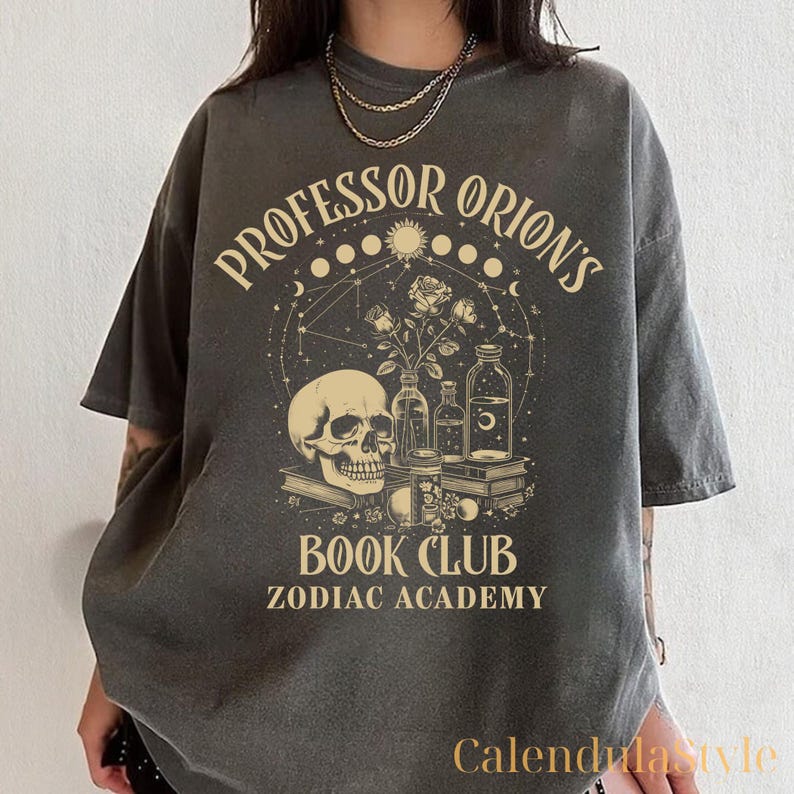 Puede incluir: Una camiseta gris oscuro con un dise&ntilde;o gr&aacute;fico que presenta una calavera, rosas y botellas. El texto "Professor Orion's Book Club Zodiac Academy" est&aacute; impreso en la camiseta.