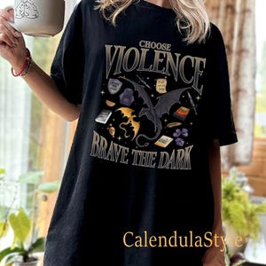 May include: Black t-shirt featuring a graphic print of a black dragon with gold accents. The text "Choose Violence Brave the Dark" is printed on the shirt.