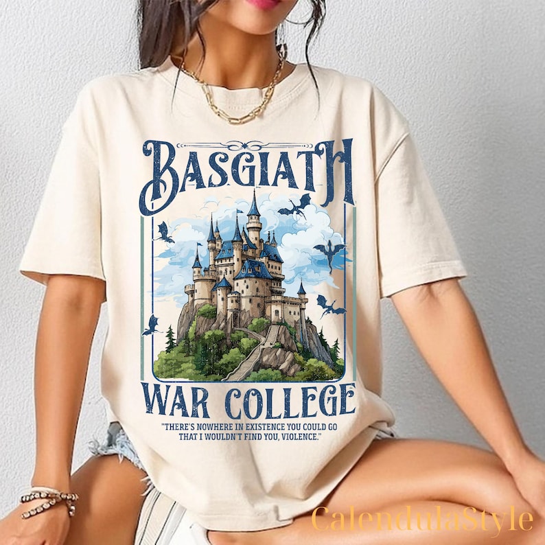 Op de afbeelding: Een beige T-shirt met een grafische print van een kasteel met draken die eromheen vliegen. De tekst "BASGIATH WAR COLLEGE" is op het T-shirt gedrukt. De quote "THERE'S NOWHERE IN EXISTENCE YOU COULD GO THAT I WOULDN'T FIND YOU, VIOLENCE." is onder de afbeelding gedrukt.