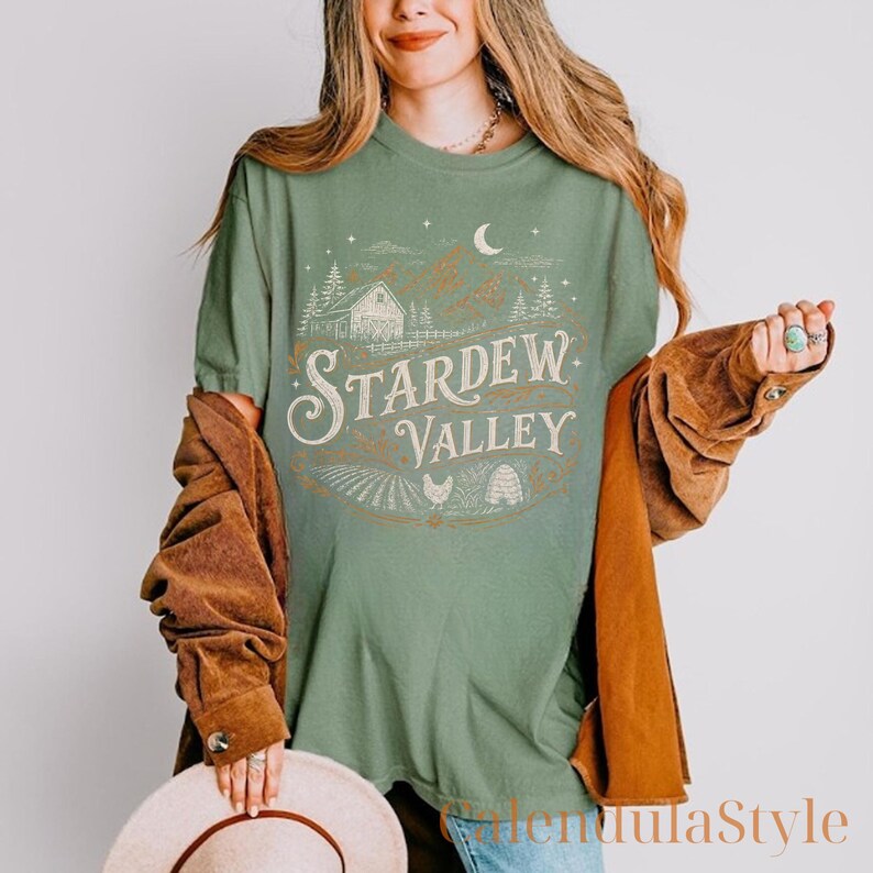 Puede incluir: Camiseta verde salvia con un dise&ntilde;o vintage con las palabras "Stardew Valley". El gr&aacute;fico incluye un granero, campos y una luna creciente. La camiseta tiene un corte holgado.