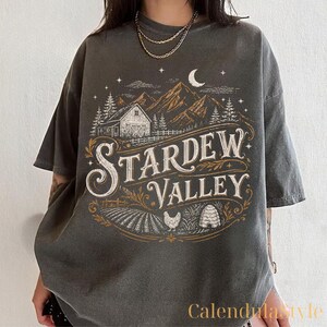 Puede incluir: Camiseta gris oscuro con un gr&aacute;fico desgastado con el texto "Stardew Valley". El dise&ntilde;o incluye un granero, monta&ntilde;as, &aacute;rboles, una luna creciente y un campo con una gallina y una colmena. La camiseta tiene mangas cortas.