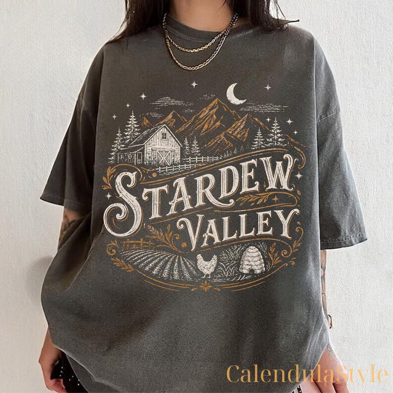 Puede incluir: Camiseta gris oscuro con un gr&aacute;fico desgastado con el texto "Stardew Valley". El dise&ntilde;o incluye un granero, monta&ntilde;as, &aacute;rboles, una luna creciente y un campo con una gallina y una colmena. La camiseta tiene mangas cortas.