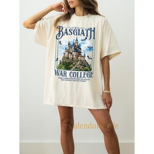 Op de afbeelding: Een cr&egrave;mekleurig T-shirt met een grafische print van een kasteel met draken en de tekst "BASGIATH WAR COLLEGE" en de quote "THERE'S NOWHERE IN EXISTENCE YOU COULD GO THAT I WOULDN'T FIND YOU, VIOLENCE."