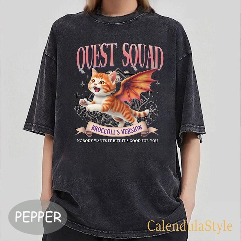 Może przedstawiać: Czarny T-shirt z grafiką kota z skrzydłami. Tekst na koszulce brzmi "Quest Squad Broccoli's Version Nobody wants it but it's good for you."