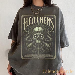 May include: Dark gray t-shirt featuring a detailed graphic with the text "HEATHENS". The design includes a skull, snakes, and a cross, with additional text below. The shirt has short sleeves and a relaxed fit.