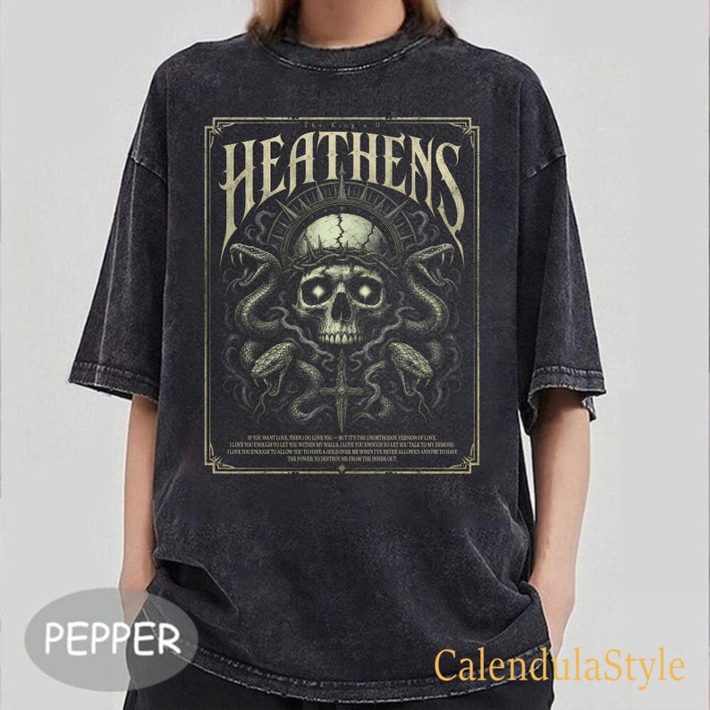 May include: Dark gray t-shirt featuring a graphic design with the text "HEATENS" above a skull with a crown and snakes. The design includes a cross and additional text at the bottom. The shirt has short sleeves.