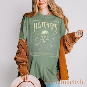 May include: Sage green t-shirt with a rectangular graphic featuring a skull, snakes, and the word "HEATTHENS" in a vintage font. The shirt is a relaxed fit and has short sleeves.