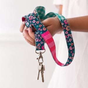 Puede incluir: Un cordón de tela con fondo verde azulado oscuro con un estampado floral rosa y naranja, una sección de rayas rosa brillante y una sección de cuadros vichy verde azulado y blanco. Incluye un mosquetón giratorio de metal plateado con dos llaves.