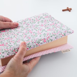 Puede incluir: Una funda de libro con estampado floral en rosa y verde sobre fondo blanco. El libro descansa sobre un cojín a rayas rosas y blancas. Una pequeña cruz de madera es visible en el fondo.