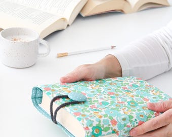 May include: A book cover with a floral pattern in shades of blue, green, and pink. The cover has a black ribbon bookmark and a blue button with white polka dots.