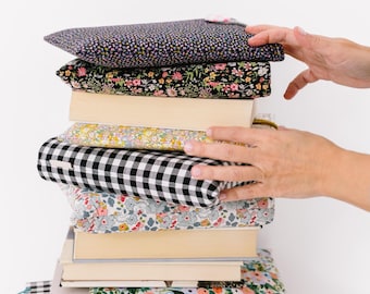 May include: A stack of books with colorful fabric book covers. The covers feature floral, polka dot, and gingham patterns in various colors. A hand is reaching towards the stack. The books are stacked on top of each other.
