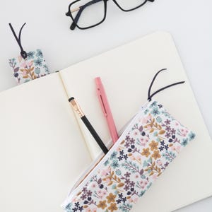May include: A floral print pencil case, bookmark, and notebook. The pencil case and bookmark feature a white background with pink, blue, and brown flowers. A black pencil and pink pen are inside the case. Black eyeglasses rest on the open notebook.