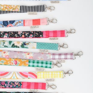 Puede incluir: Una colección de doce cordones de tela con varios patrones que incluyen flores, cuadros vichy, rayas y lunares. Cada cordón tiene un mosquetón giratorio de metal plateado y una pequeña etiqueta de tela blanca con el texto DRAPETS.