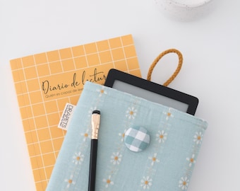 May include: A light blue e-reader case with a daisy pattern and a gingham button, holding a black e-reader. A yellow grid notebook with the text "Diario de lectura" and a black pen with a gold tip are also present. A cup of coffee is in the background.