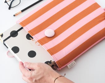 May include: A pink and orange striped notebook sleeve with a matching button closure. Inside the sleeve is a spiral-bound notebook with black polka dots. A pair of black glasses and a pencil are on the white surface.
