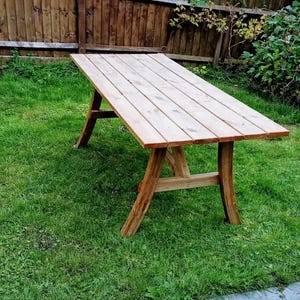 May include: A light brown wooden table with a rectangular top and angled legs, set on a green lawn. The table is approximately 152 cm long, with a wooden fence and greenery in the background. Ideal for outdoor use.