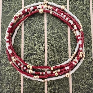 May include: A collection of beaded bracelets arranged in a circle. The bracelets are made with red, white, and gold beads. The background is a green surface with white lines and numbers, similar to a football pitch.