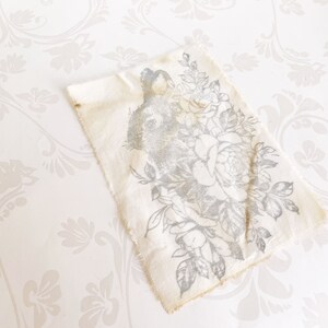 May include: A white fabric square with a gray floral design featuring a rabbit surrounded by roses. The fabric has a frayed edge.