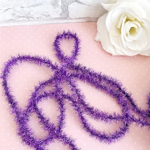May include: A close-up of a length of purple tinsel, with a loop at the top. The tinsel is a vibrant, shimmering purple color. A white rose is in the background, with a pink polka dot background.