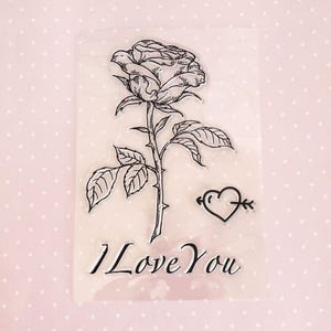 May include: Clear stamp sheet featuring a black line drawing of a rose with thorns, a heart with an arrow through it, and the text "I Love You".