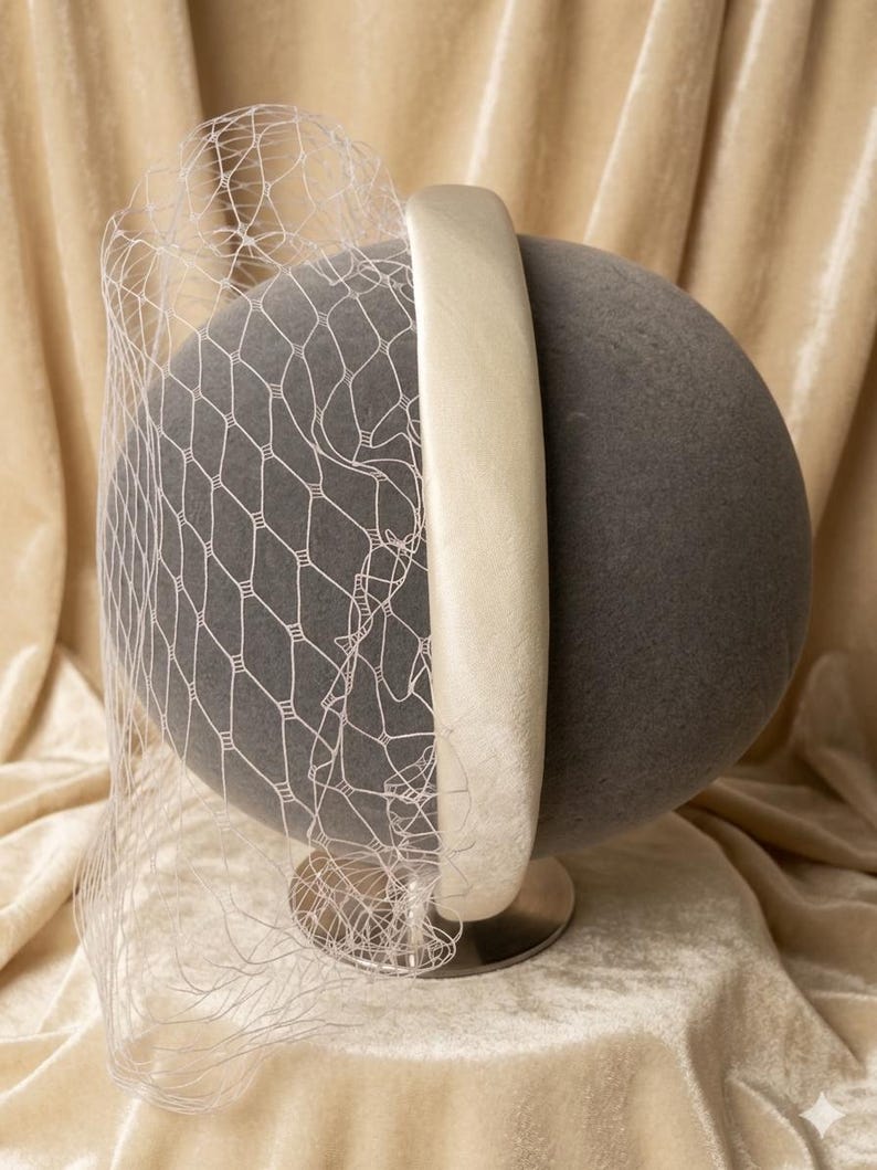 Op de afbeelding: Een grijze, bolvormige hoed met een cr&egrave;mekleurige hoofdband en een witte netsluier. De hoed staat op een zilverkleurige basis, tegen een achtergrond van zachte, cr&egrave;mekleurige stof. De sluier drapeert over de voorkant van de hoed.