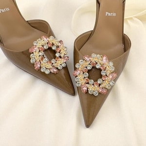 May include: A pair of brown high-heeled shoes with pointed toes. Each shoe features a decorative, circular embellishment with pink and clear crystals and gold-colored metal accents. The word "Paris" is printed on the insole.