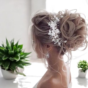Pode incluir: Um pente de cabelo floral branco com detalhes em pérolas. O pente é usado no cabelo de uma mulher, que está penteado em um coque desarrumado.