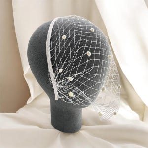 May include: A white birdcage veil attached to a thin headband, featuring a delicate diamond-shaped netting pattern adorned with small, round, off-white faux pearls scattered across the surface, creating a textured and layered appearance.