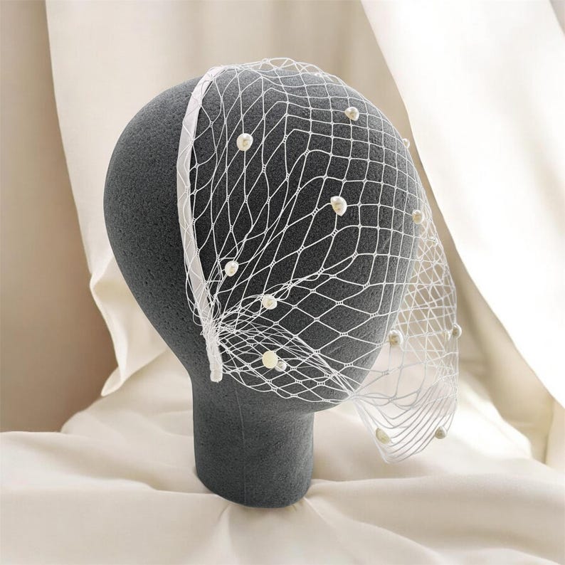 May include: A white birdcage veil attached to a thin headband, featuring a delicate diamond-shaped netting pattern adorned with small, round, off-white faux pearls scattered across the surface, creating a textured and layered appearance.