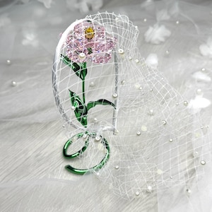 May include: A white headband featuring a pink crystal flower with a yellow centre, green metal stem and leaves, and a white netting veil adorned with small white pearls. The base of the stem forms a circular green metal loop for stability.